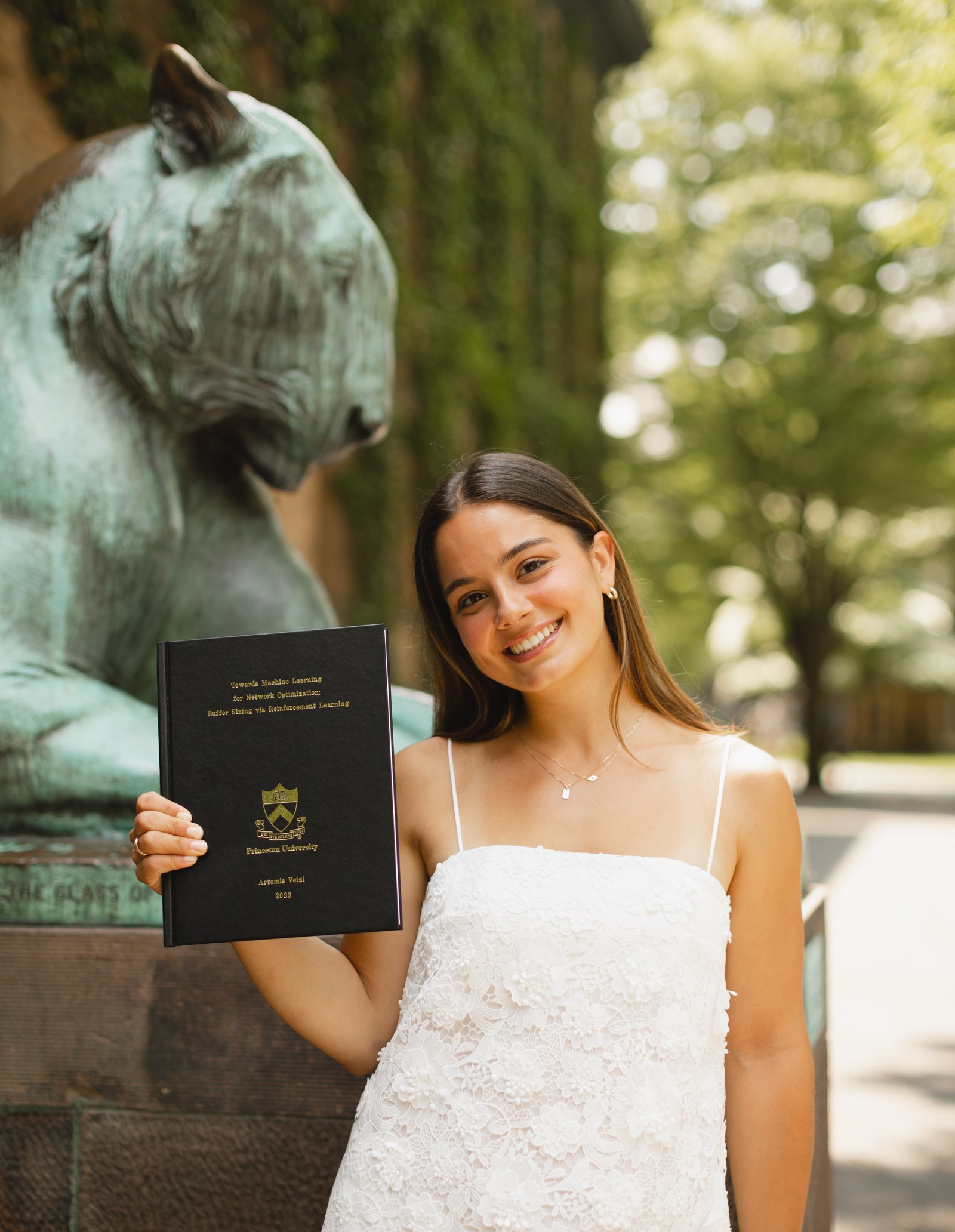 Artemis Veizi holding her bound Princeton senior thesis, &ldquo;Towards Machine Learning for Network Optimization: Buffer Sizing via Reinforcement Learning&rdquo;, beside the Princeton tiger statue.